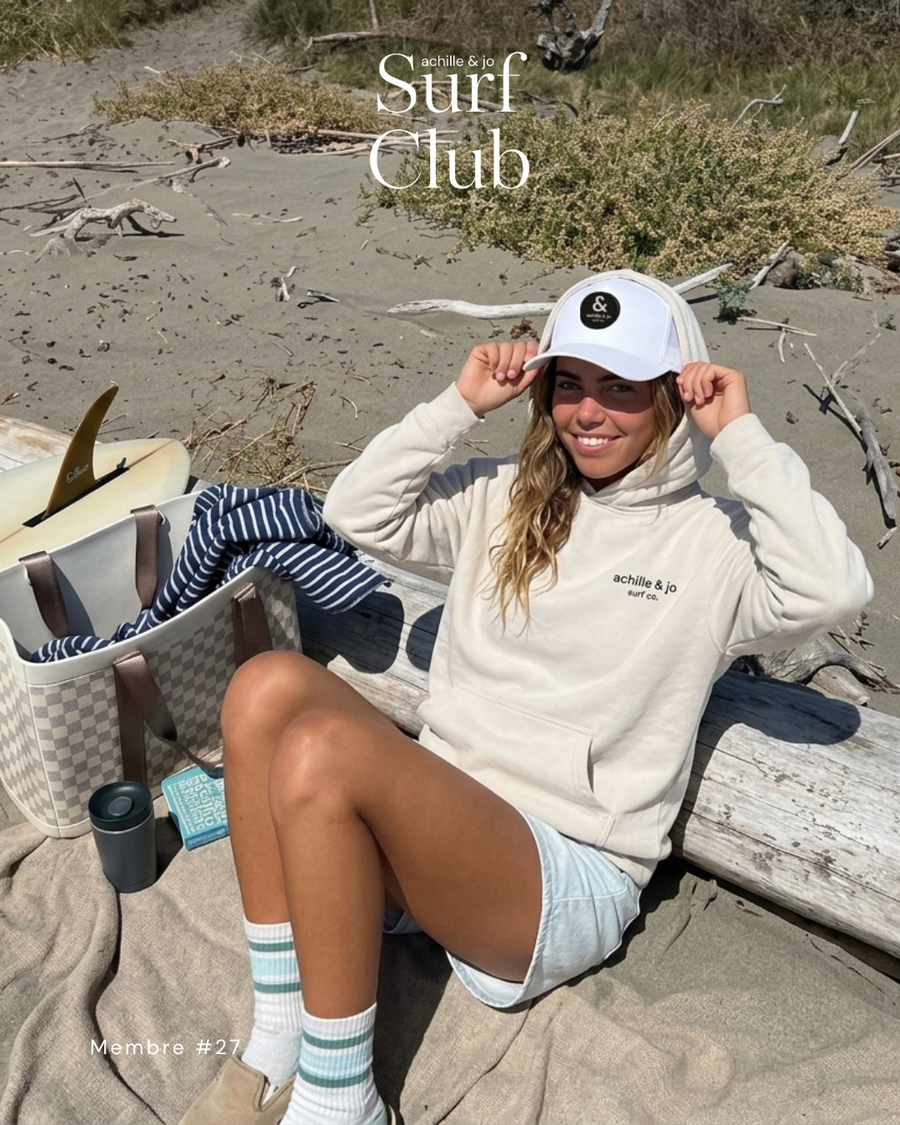 Membre du Surf Club Achille & Jo en hoodie écru et casquette à patch, sur la plage